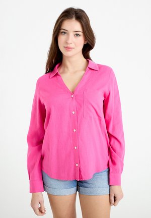 Junge Frau trägt ein knallpinkes Hemd mit Knöpfen und hellblaue, umgekrempelte Jeansshorts vor weißem Hintergrund.