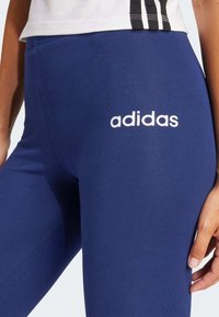 Blå sportleggings gjorda av stretchigt material, med en vit "adidas"-logotyp på låret och en slät yta. Inga synliga sömmar.