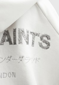 Witte sweatshirt met zilverkleurige pailletten tekst: "SAINTS" en Japanse karakters in een vet, modern lettertype. Zacht, gestructureerd stof.
