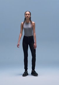 Szürke ujjatlan body, fekete karcsúsított farmernadrág, fekete fűzős cipő. Semleges testtartásban áll világoskék háttér előtt.