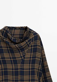 Blouse à manches longues à carreaux bleu marine et beige avec col bénitier, posée à plat sur un fond blanc.