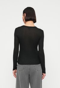 Γυναίκα με ίσια σκοτεινά μαλλιά, ντυμένη με εφαρμοστή μαύρη μπλούζα μακρυμάνικη και ψηλόμεσα γκρι τζιν, εμφανιζόμενη από πίσω σε απλό φόντο.