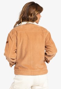 Vrouw met golvend bruin haar, gekleed in een tan corduroy jas met een witte schapenvacht kraag, met haar rug naar de camera gekeerd tegen een effen witte achtergrond.
