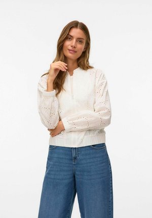 White bomber jacket with textured, embroidered detailing and elastic cuffs, paired with loose, light blue jeans. Simple zip closure at front.