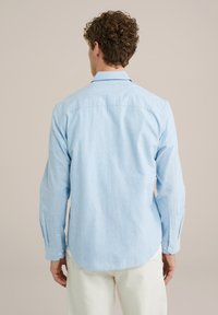 Lichtblauw overhemd met lange mouwen, gemaakt van een gladde stof. Beschikt over een button-down kraag, zijplooien en knopen op de manchetten. Achteraanzicht.