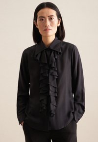Blusa nera con un grande dettaglio anteriore a volant, maniche lunghe e colletto a punta. Tessuto morbido e vestibilità sagomata. Polsini neri semplici.