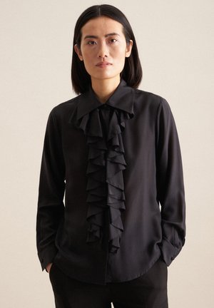 Blusa nera con un grande dettaglio anteriore a volant, maniche lunghe e colletto a punta. Tessuto morbido e vestibilità sagomata. Polsini neri semplici.