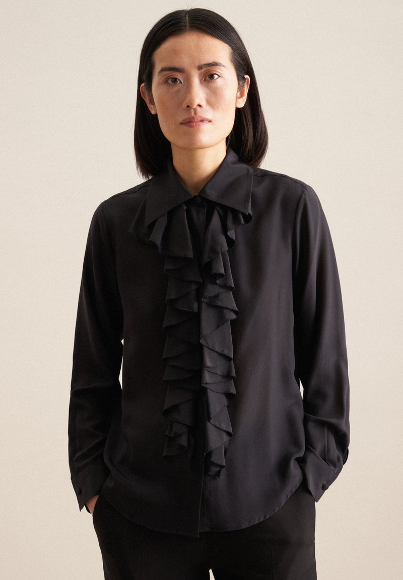 Blusa nera con un grande dettaglio anteriore a volant, maniche lunghe e colletto a punta. Tessuto morbido e vestibilità sagomata. Polsini neri semplici.