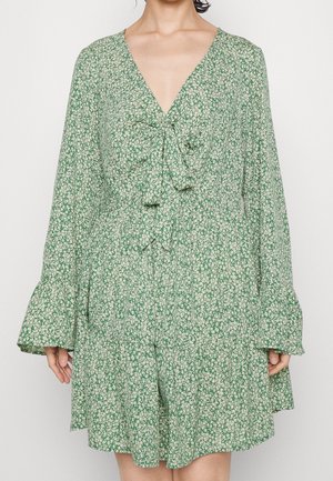 Robe verte avec un petit imprimé floral blanc, manches longues évasées, col en V et un nœud à l'avant à la taille.