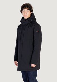 Giacca isolante nera con colletto alto, tasche frontali e cappuccio con cordino regolabile. Presenta discreti accenti con il marchio.