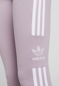 Světle fialové legíny z jemné tkaniny mají bílé tři pruhy Adidas a logo vyšitý blízko dolní části nohy.