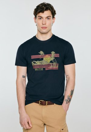 Młody mężczyzna z kręconymi włosami, ubrany w granatowy t-shirt z napisem „BIG STAR” i beżowe spodnie, stoi na jednolitym białym tle.