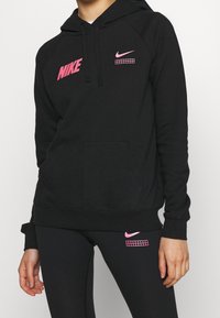 Svart huvtröja med rosa Nike-logotyper på bröstet och ärmen, med en kenguruficka och ribbade muddar, tillverkad av ett mjukt material.