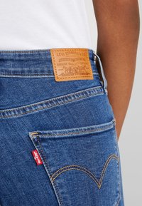 Jeans de mezclilla azul con una etiqueta de cuero marrón claro en la parte trasera con texto en relieve y costuras. Etiqueta roja con logo en el bolsillo trasero, detalle de costura curvada.