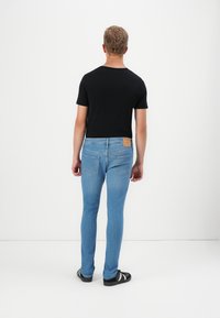 Jeans skinny de denim azul con un lavado claro, dos bolsillos traseros y un parche de cuero marrón en la cintura. Camiseta negra y zapatos negros.
