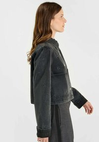 Chaqueta de denim negra con un diseño recortado, que presenta un cierre de botón, cuello puntiagudo y patrones sutiles de logotipos a lo largo de la tela.