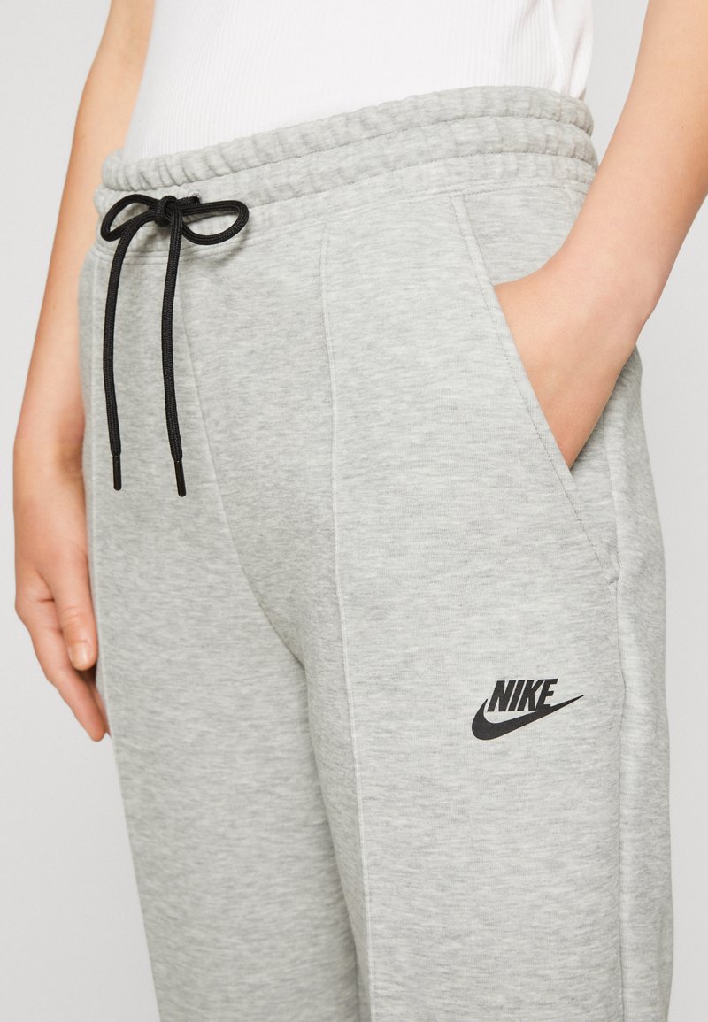 Pantalones de chándal grises con cintura ajustable, bolsillos laterales y un logo de Nike negro en el muslo izquierdo. Tejido suave y texturizado.