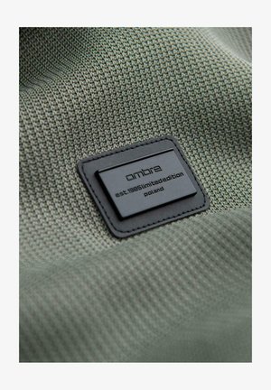 Etichetta rettangolare nera Ombré del marchio su tessuto verde testurizzato con la scritta "est. 1985 edizione limitata Polonia".