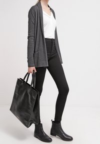 Cárdigan gris con bolsillos, blusa blanca de capas, jeans ajustados negros de cintura alta, botines de cuero negros y un gran bolso negro.