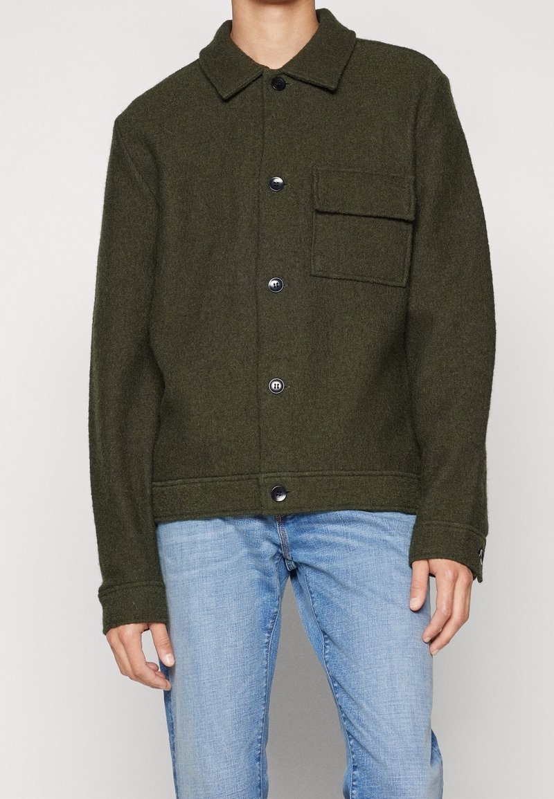 Grön jacka i ullblandning med knäppning fram, standard krage och bröstficka; kombinerad med ljusblå denimjeans.