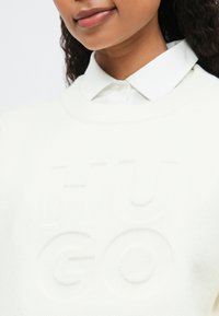 Νέος άνθρωπος που φοράει ένα λευκό πουκάμισο με κολάρο κάτω από ένα λευκό πουλόβερ με διακριτικό ανάγλυφο κείμενο "LEGO", χαμογελώντας ελαφρώς, μπροστά από απλό φόντο.