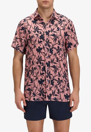 Uomo che indossa una camicia rosa a maniche corte con motivo floreale nero e pantaloncini blu navy, in piedi con le braccia rilassate lungo i fianchi.