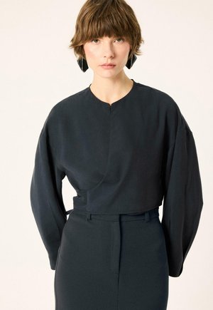 Jeune personne aux cheveux courts et bruns portant un haut noir ample à manches longues et un pantalon taille haute assorti sur un fond uni.