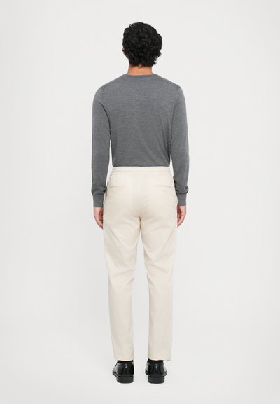 Cămașă lungă gri deschis cu mâneci lungi, pantaloni de culoare crem cu un brâu elastic, și pantofi negri, văzuți din spate pe un fundal simplu.