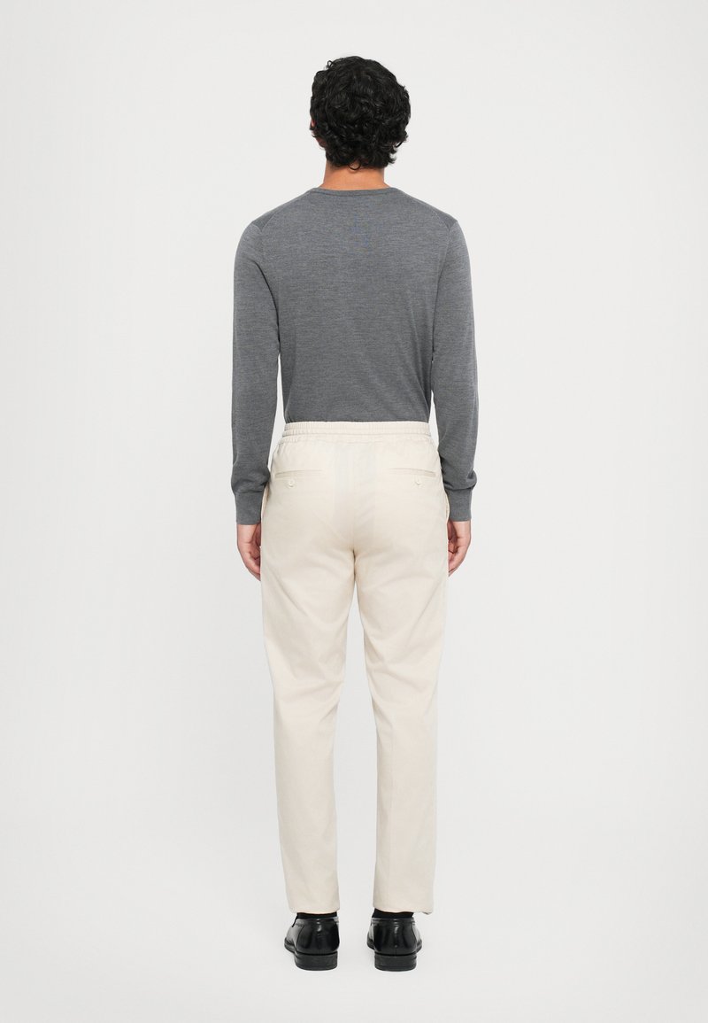 Lichtgrijs langemouwen shirt, crèmekleurige broek met een elastische tailleband, en zwarte schoenen, gezien vanuit de achterkant tegen een effen achtergrond.