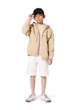 Beige leichte Jacke mit zwei Fronttaschen, cremefarbener Pullover, weiße Shorts und grau-weiße Sneaker. Schwarze Kappe mit Logo.