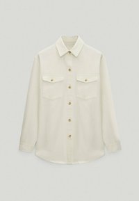 Chemise beige à manches longues avec col pointu et deux poches poitrine à rabats boutonnés.