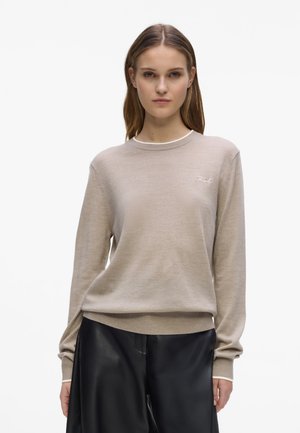 Femme portant un pull beige à col rond avec un petit logo blanc et un pantalon noir à jambes larges, debout sur un fond blanc uni.