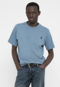 Φωτεινό γαλάζιο κοντομάνικο t-shirt με στρογγυλή λαιμόκοψη, που διαθέτει τσέπη στο στήθος και διακριτική υφή, συνδυασμένο με σκούρο τζιν και μαύρη ζώνη.