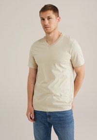 Beige v-veida t-krekls, izgatavots no mīksta auduma, ar īsām piedurknēm un brīvu piegriezumu, kombinēts ar ziliem džinsiem, kurus valkā stāvošs vīrietis.