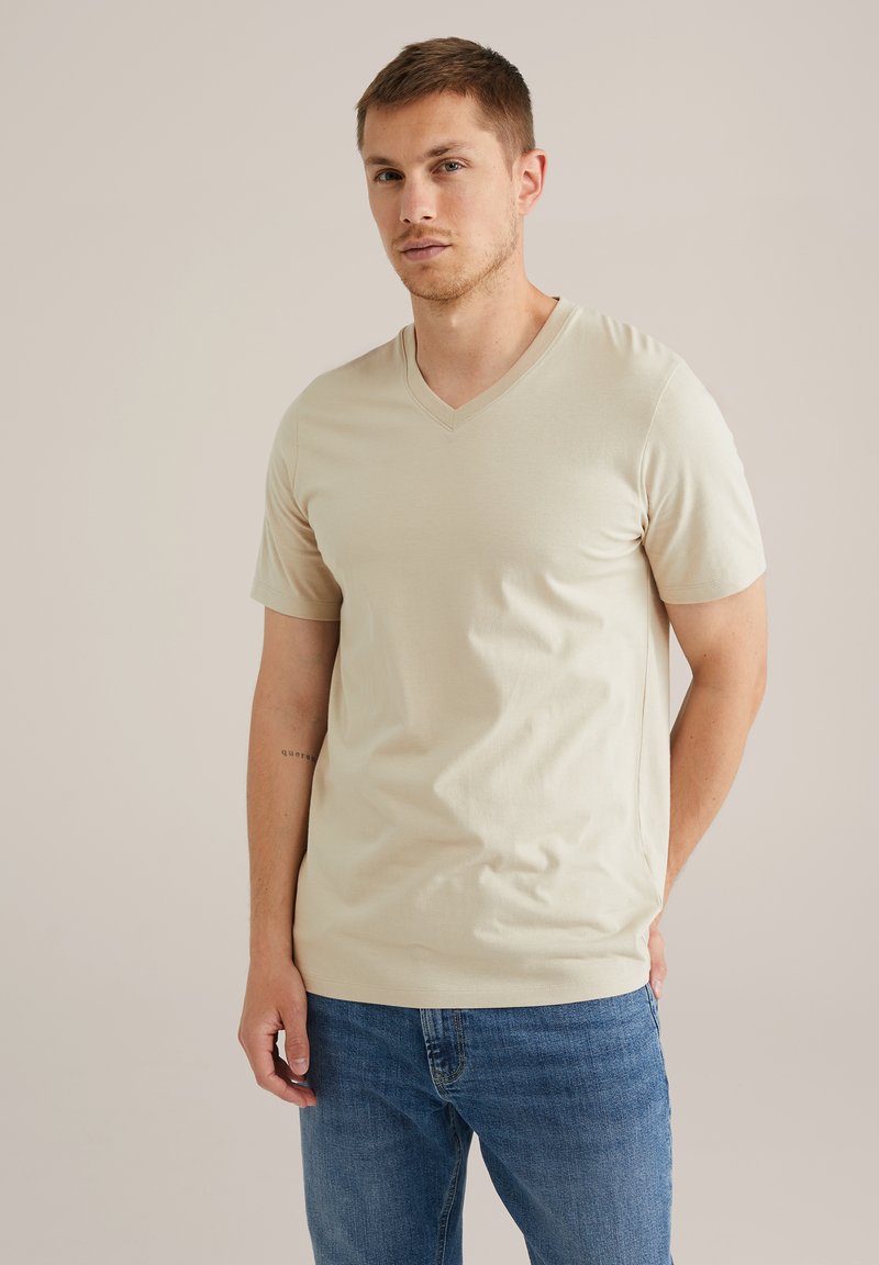 Beige v-veida t-krekls, izgatavots no mīksta auduma, ar īsām piedurknēm un brīvu piegriezumu, kombinēts ar ziliem džinsiem, kurus valkā stāvošs vīrietis.