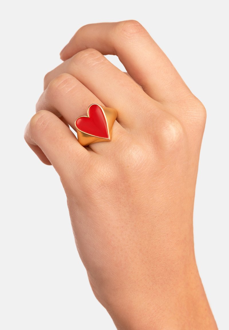 Anillo de metal dorado con un gran diseño de corazón rojo, acabado suave y brillante, llevado en una mano con textura de piel visible.