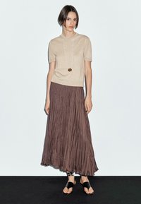 Un modello è in piedi su uno sfondo chiaro, indossando un top beige e una gonna marrone fluente, completato da sandali neri con cinturini e una collana.