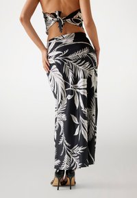 Fustă maxi neagră cu un model de frunze tropicale albe, având un tiv suprapus, dintr-un material neted, purtată cu pantofi cu toc negri.