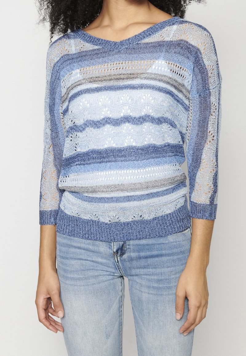 Pull tricoté à rayures bleues avec manches trois-quarts, présentant un motif en dentelle et des poignets texturés. Porté avec un jean en denim clair.