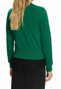 Sudadera de algodón verde con dobladillo y puños acanalados, que presenta un diseño sencillo de cuello redondo. Llevada sobre una blusa negra en capas.