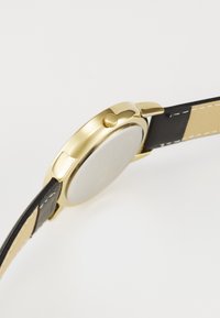 Montre dorée avec un boîtier rond, un bracelet en cuir noir et un cadran blanc. Comprend une couronne sur le côté pour les réglages. Texture lisse.