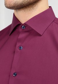 Chemise à manches longues en tissu bordeaux profond. Elle présente un col pointu et des boutons bleus, avec une texture lisse et sans motifs visibles.