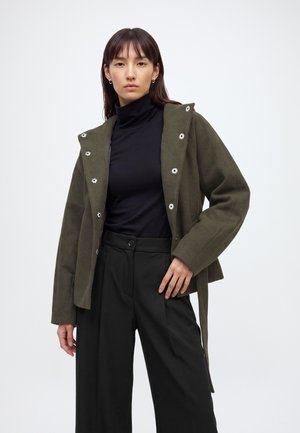Femme portant une veste verte olive ouverte sur un col roulé noir et un pantalon noir taille haute sur un fond blanc uni.