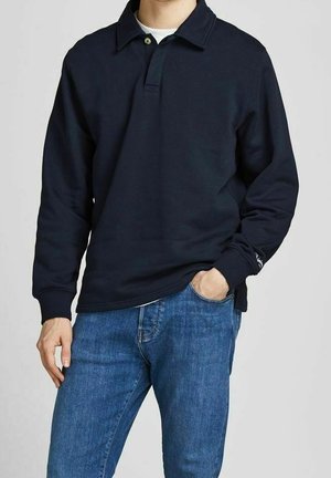 Mand iført sort langærmet poloshirt over hvid undertrøje og blå jeans, den ene hånd i lommen, mod en ensfarvet baggrund.