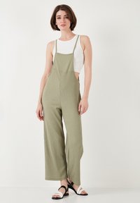 Salopette verde oliva a gamba larga con bretelle regolabili e tessuto texturizzato, abbinata a una canotta bianca aderente e sandali neri.