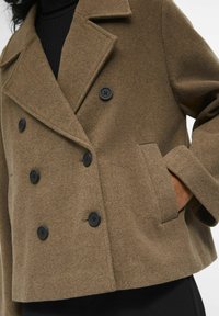 Manteau double boutonnage en mélange de laine marron avec de grands boutons noirs, revers crantés et poches latérales. Texture douce et coupe décontractée.