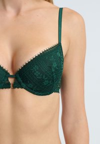 Primer plano de una mujer que lleva un sujetador de encaje verde oscuro con tirantes finos y patrones florales sobre un fondo claro y liso.