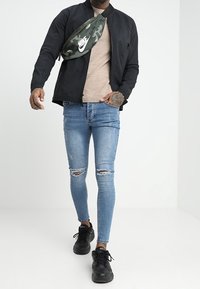 Czarna kurtka bomber, beżowa koszula, jasnoniebieskie podarte dżinsy i czarne sneakersy. Zielona torba na ramię w stylu camo z białym logo Nike.