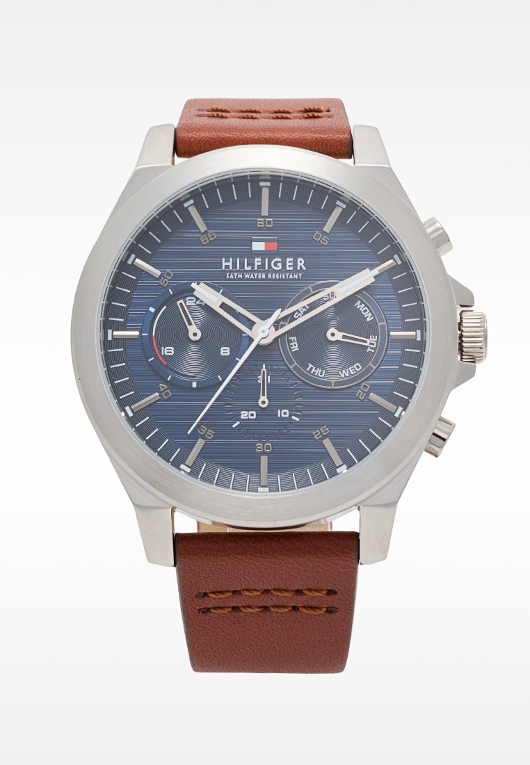 Reloj de acero inoxidable plateado con esfera azul marino, correa de cuero marrón, índices horarios elevados y subesferas para la fecha y el día. Se destaca la característica de resistencia al agua.