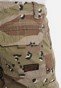 Pantalones cortos de carga de camuflaje en tonos marrón y verde con detalles en negro. Las características incluyen bolsillos, presillas para cinturón y una etiqueta de tela con detalles de marca.